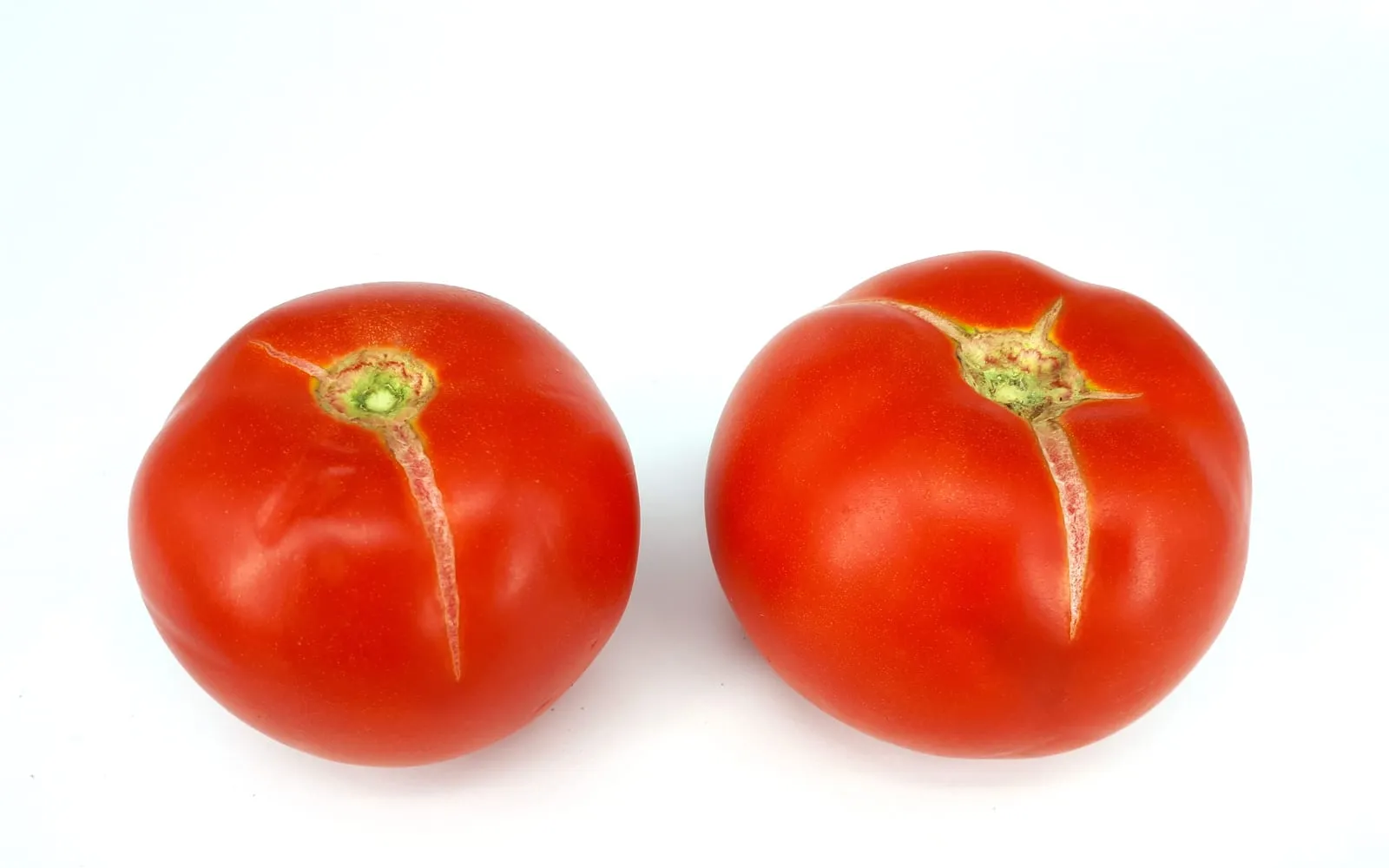 Two ripe red tomatoes photographed against white, each with a single pale healed split running from the stalk scar down the shoulder
