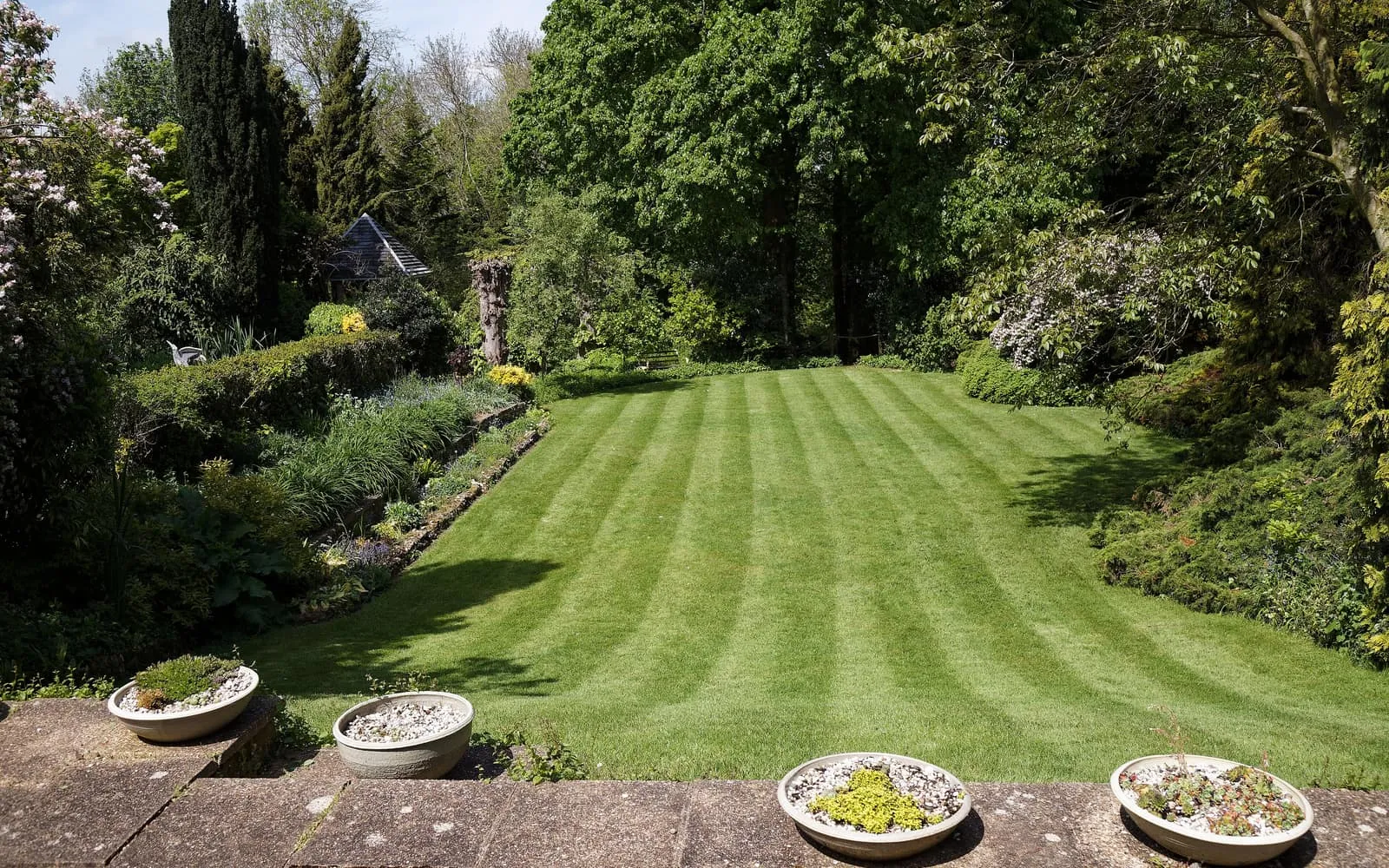 A neatly striped garden lawn framed by planted borders and mature trees, with shallow stone alpine pots on a paved terrace in the foreground