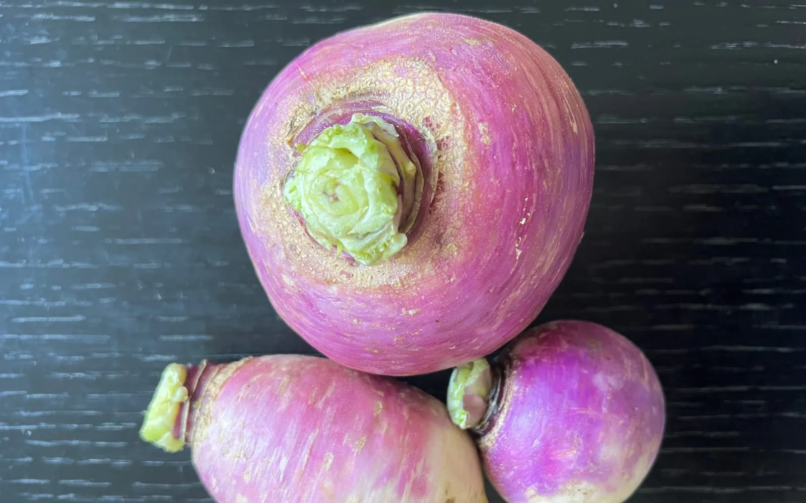 Three purple-topped white turnips on a dark wooden surface, cut leaf stalks still attached